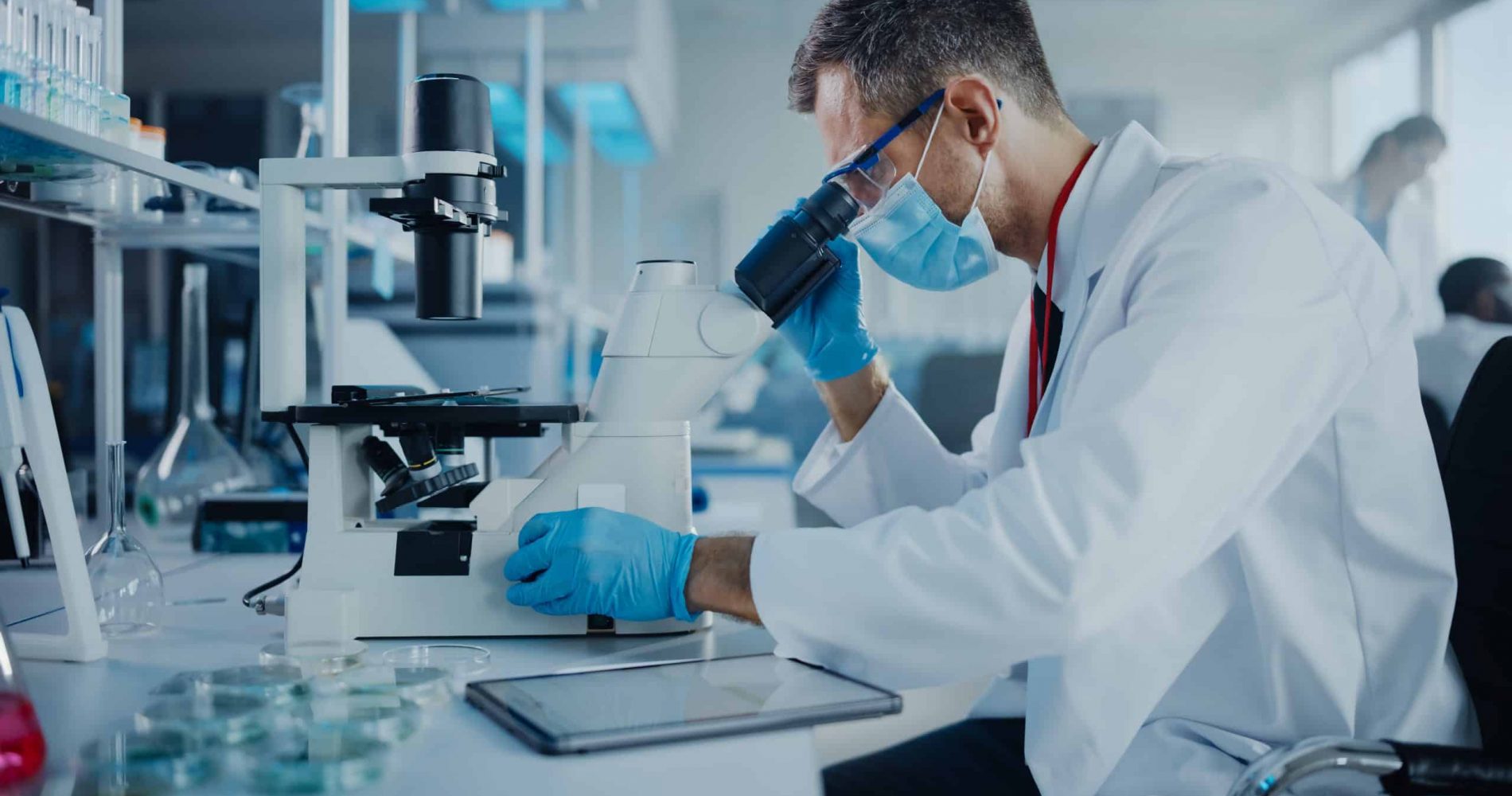 Medical Development Laboratory: Scientist Wearing Face Mask Looking Under Microscope and Using Digital Tablet. Specialists Working on Medicine, Biotechnology Research in Advanced Pharma Lab